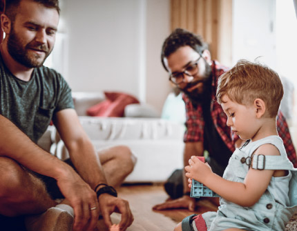 A toddler boy sitting on the floor plays with blocks while two men join him in play.